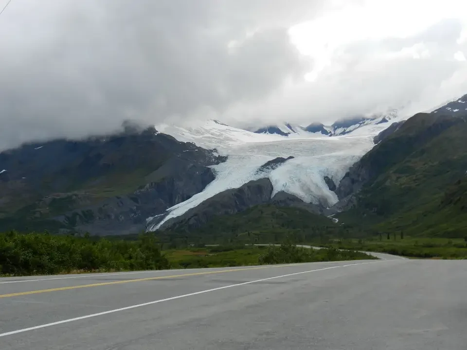 A road meanders through a mountain landscape with glaciers under an overcast sky.