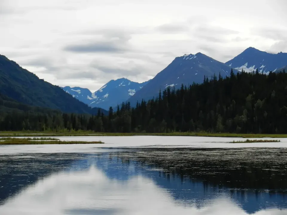 An alpine lake reflects snow-capped mountains and green forests under a cloudy sky.