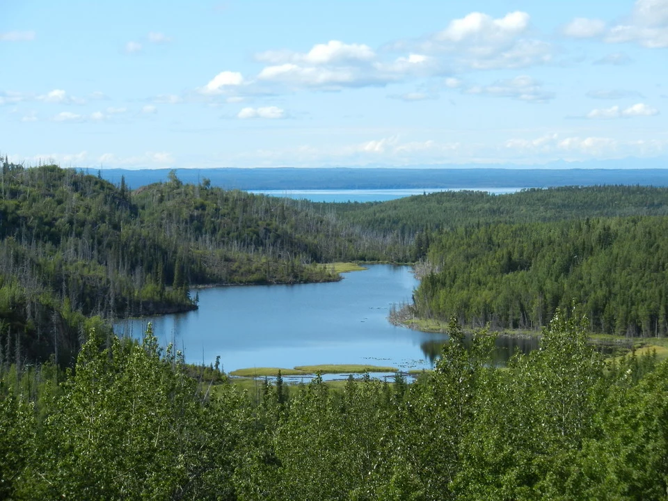 A vast landscape shows lakes surrounded by dense coniferous forest under a partly cloudy sky.