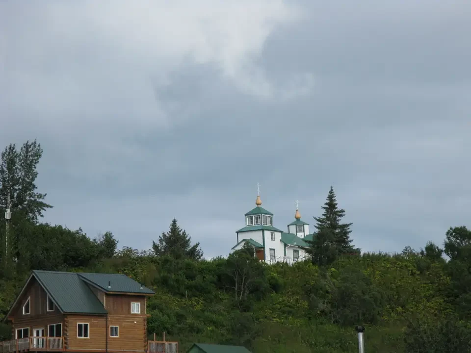 A green house with domed roofs stands on a forested hill under a cloudy sky.
