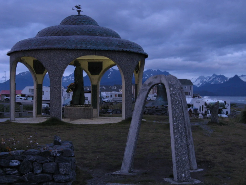 A stone structure with a domed roof sits on a grassy lot with mountains in the background.