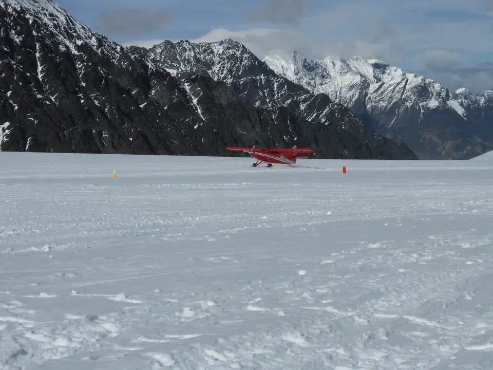 A red plane sits over a vast snowy landscape with snow-capped mountains in the background.