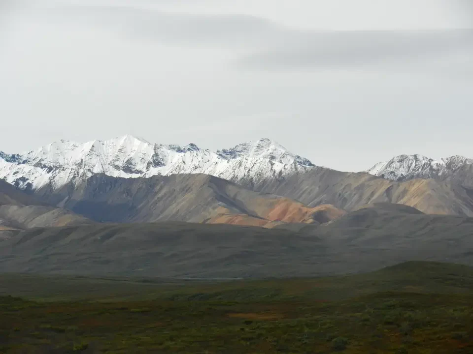 Snow-capped mountain ranges stand out against a hilly landscape under an overcast sky.