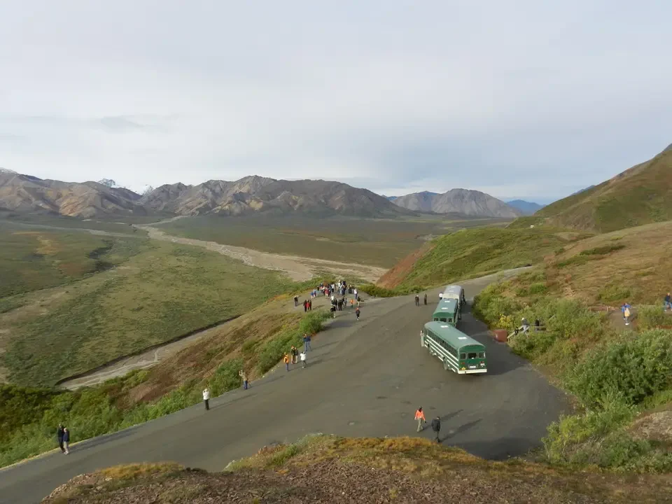 A group of people and a bus travel along a dirt road through a mountain landscape.
