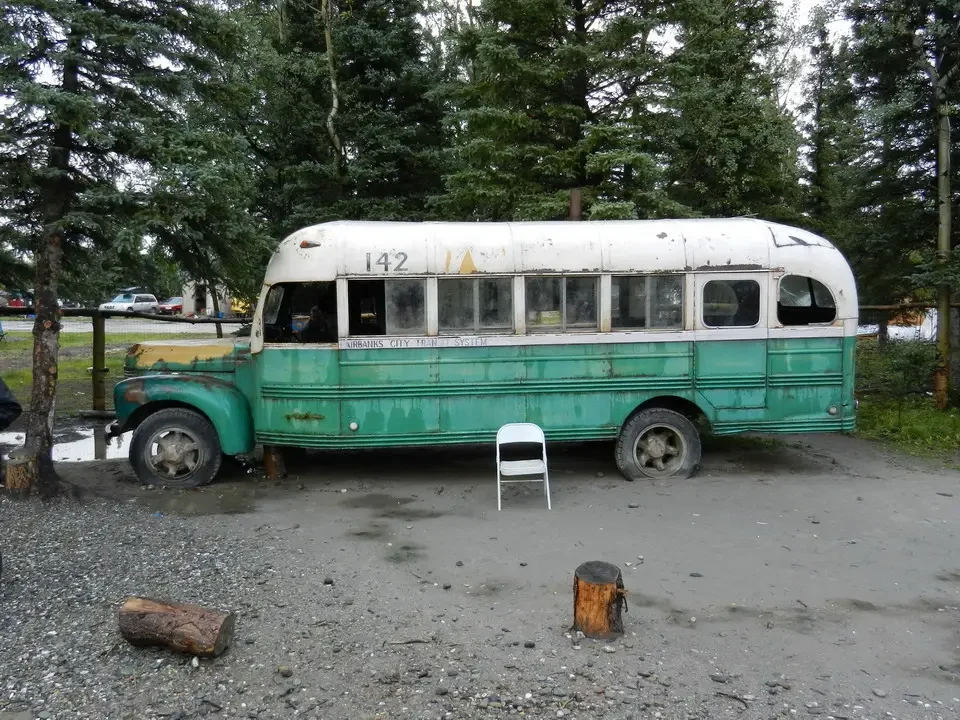 An old green and white bus is parked on a dirt lot among trees.