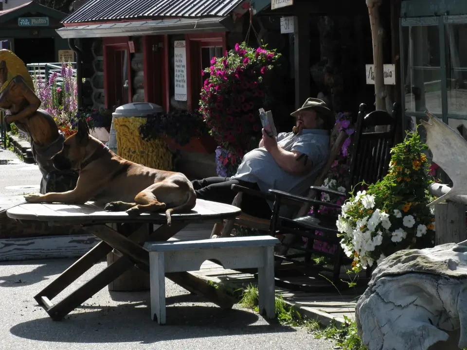 A man reads on an outdoor bench with a dog lying next to a wooden building.