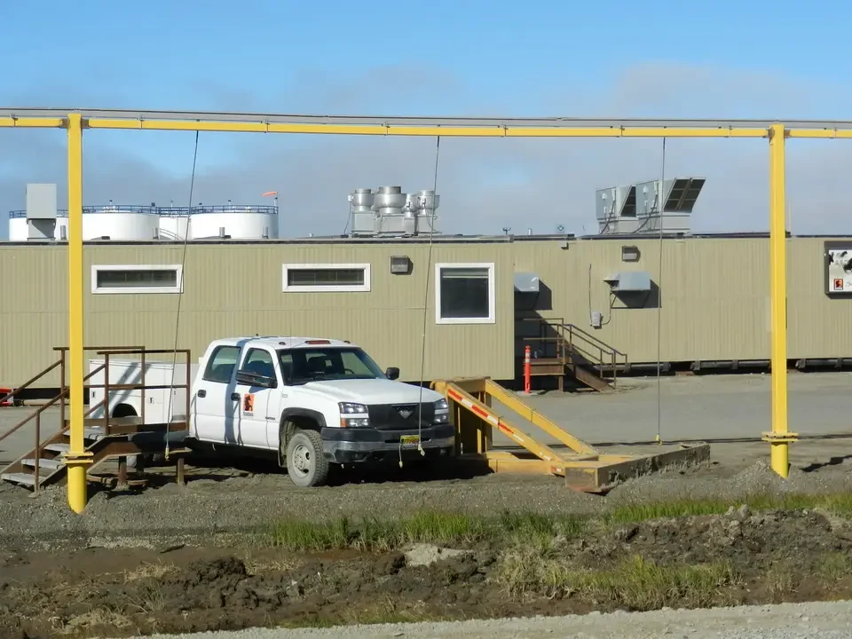 A white truck parked in front of a long building with a yellow structure above it, in an outdoor, open setting.