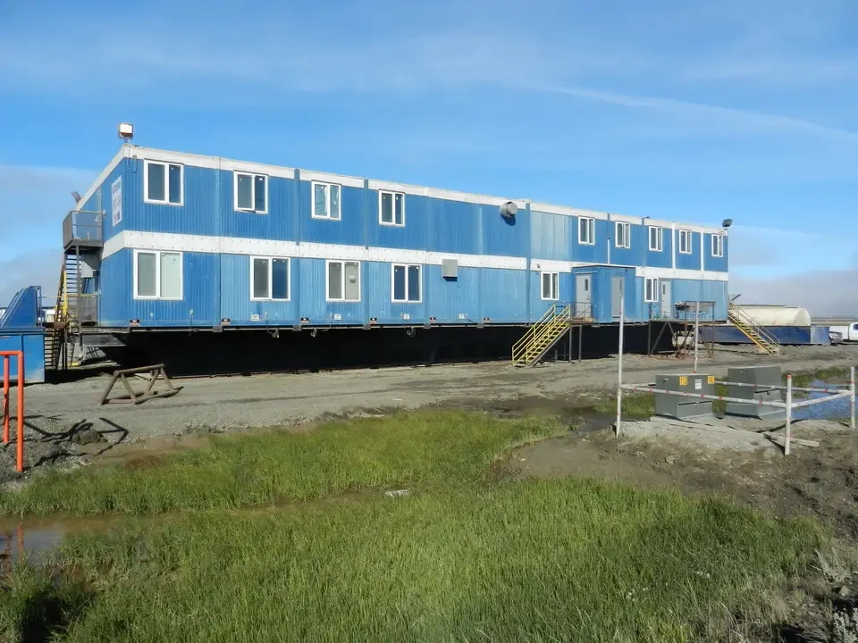 A long blue building sits on flat land with greenery in the foreground.