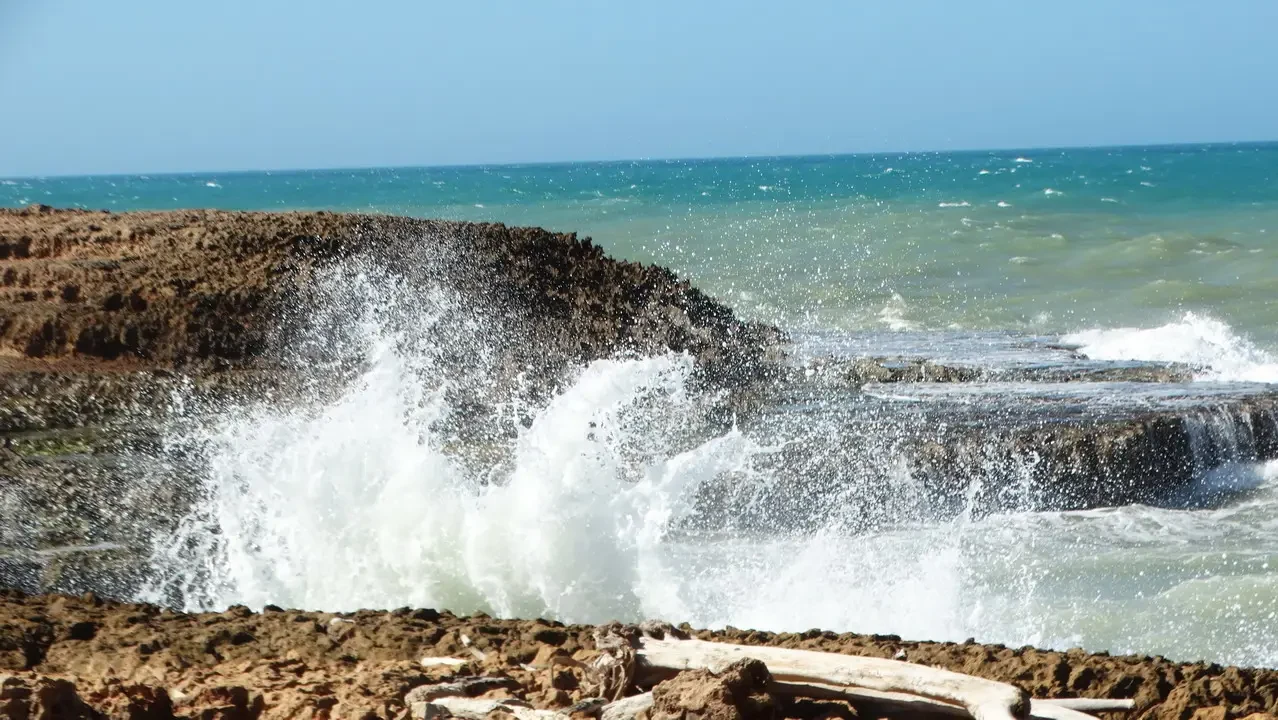 Onde d'acqua turchesi si infrangono sulla riva rocciosa con schizzi bianchi.
