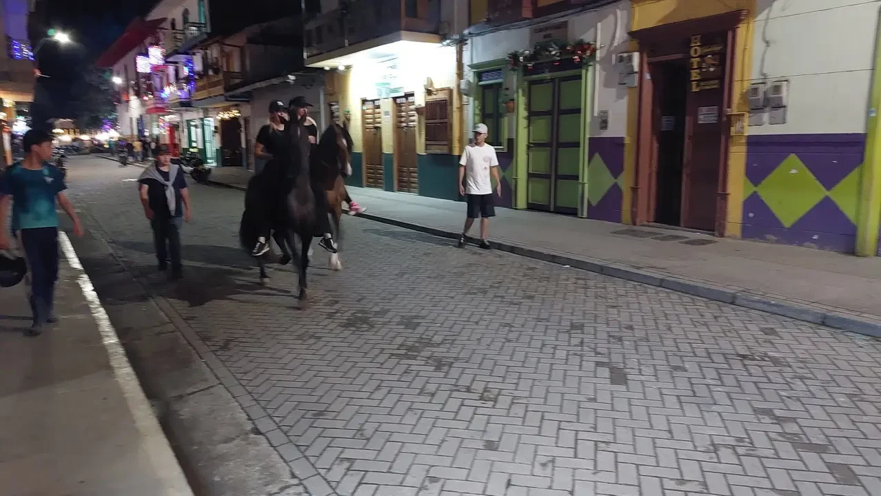 Persone e cavalli sfilano lungo una strada acciottolata in un quartiere colombiano storico.