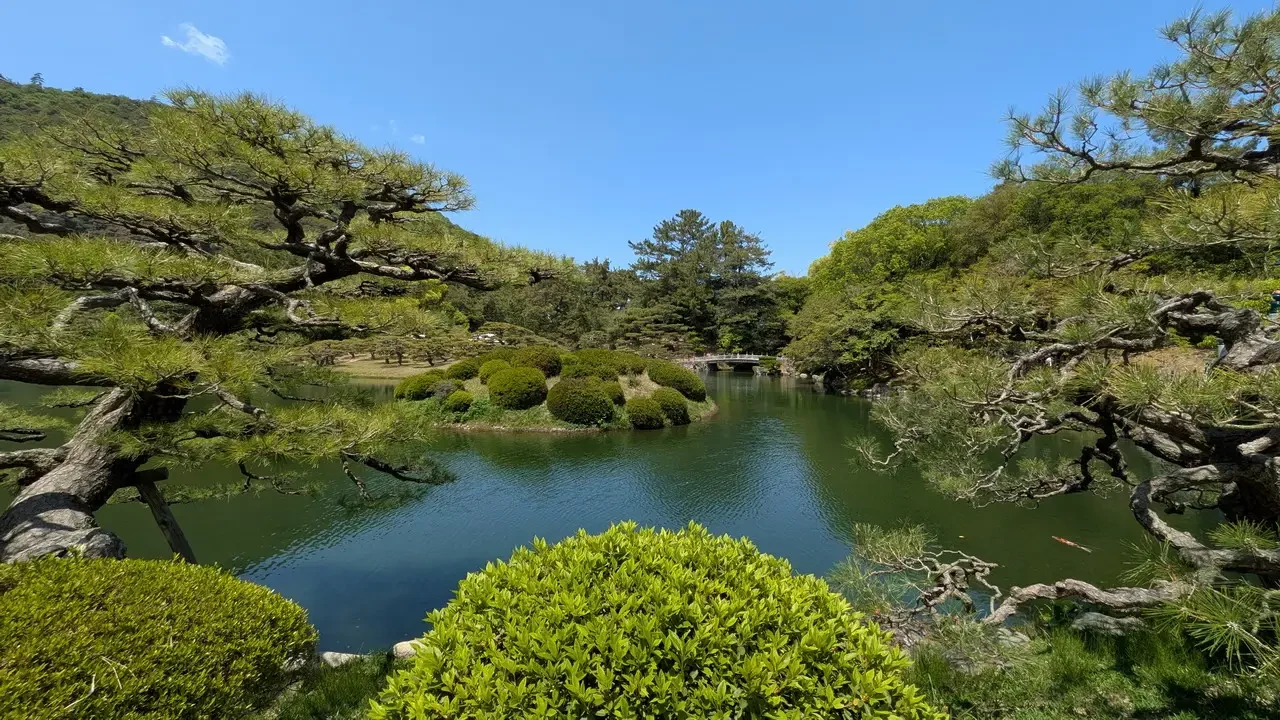 Giardino Ritsurin in Giappone con alberi e acqua verde sotto il cielo azzurro.
