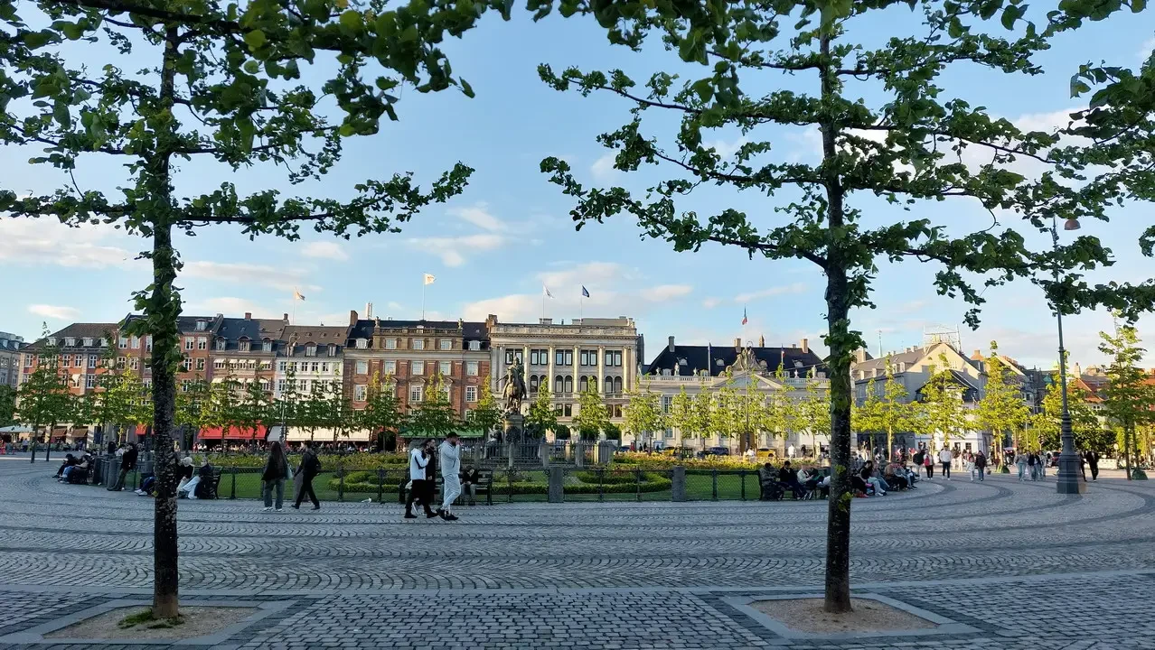 Vista di una piazza europea con alberi in primo piano e edifici storici sullo sfondo.