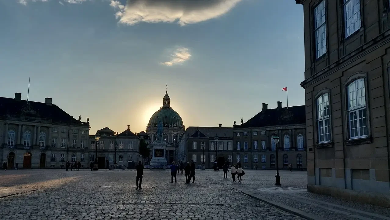 Una piazza storica europea è illuminata dalla luce del tramonto con edifici classici e persone che passeggiano.
