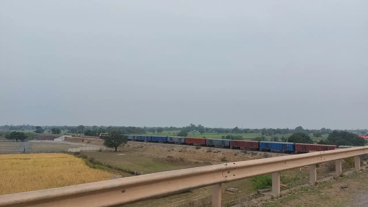 Treno merci attraversa un paesaggio rurale indiano sotto un cielo coperto.