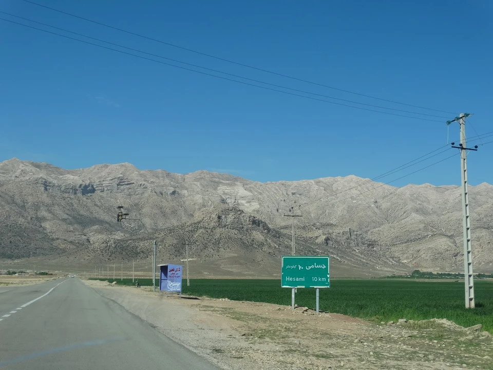 Una strada asfaltata attraversa un paesaggio montuoso in Iran con campi coltivati ai lati.