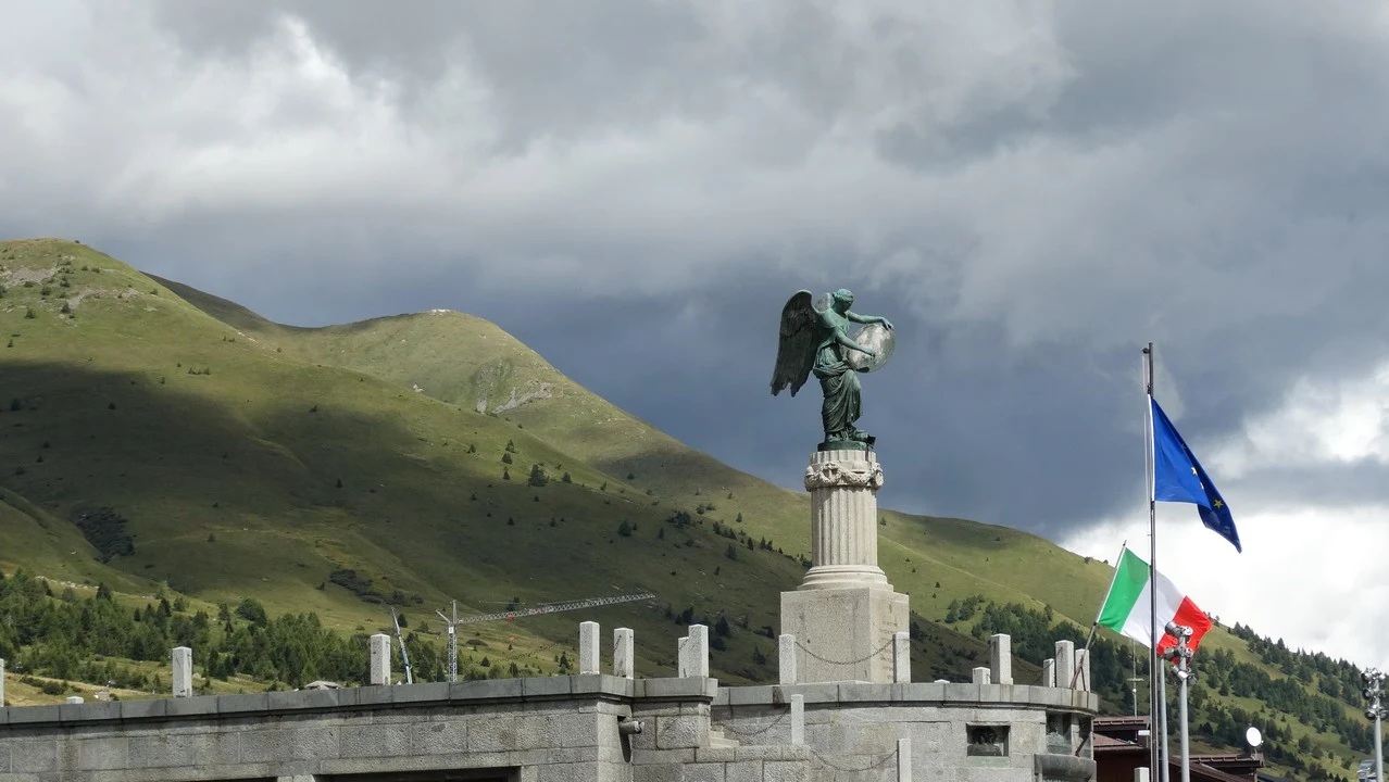 Statua angelica su piedistallo in pietra con vista sulle montagne alpine sotto un cielo nuvoloso.