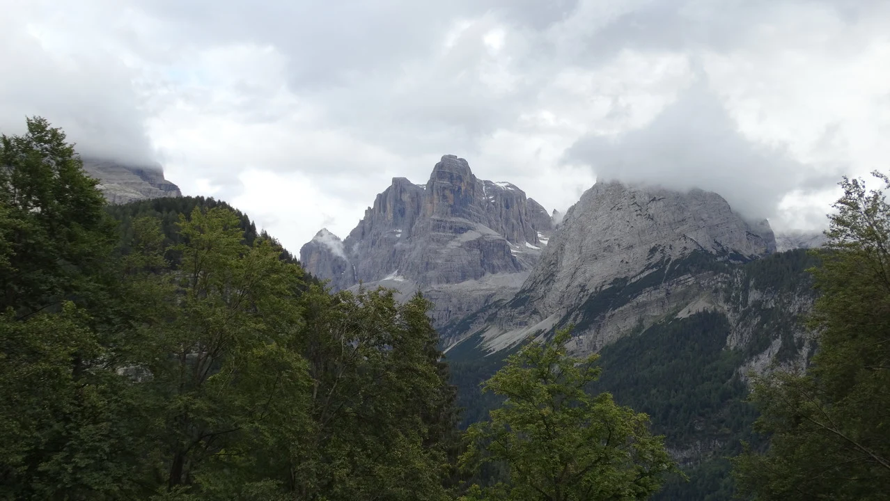 Paesaggio montano delle Alpi con cime rocciose e foreste verdi sotto un cielo nuvoloso.