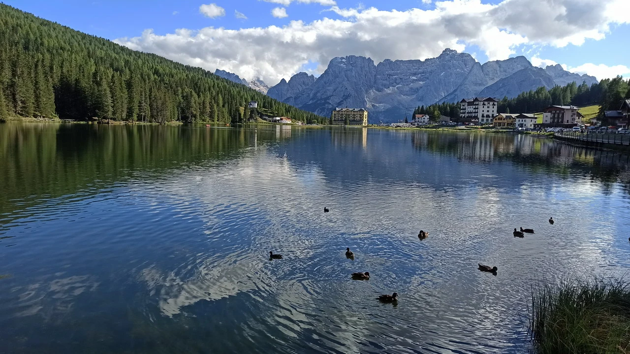 Cavalieri attraversano un lago alpino con montagne sullo sfondo.