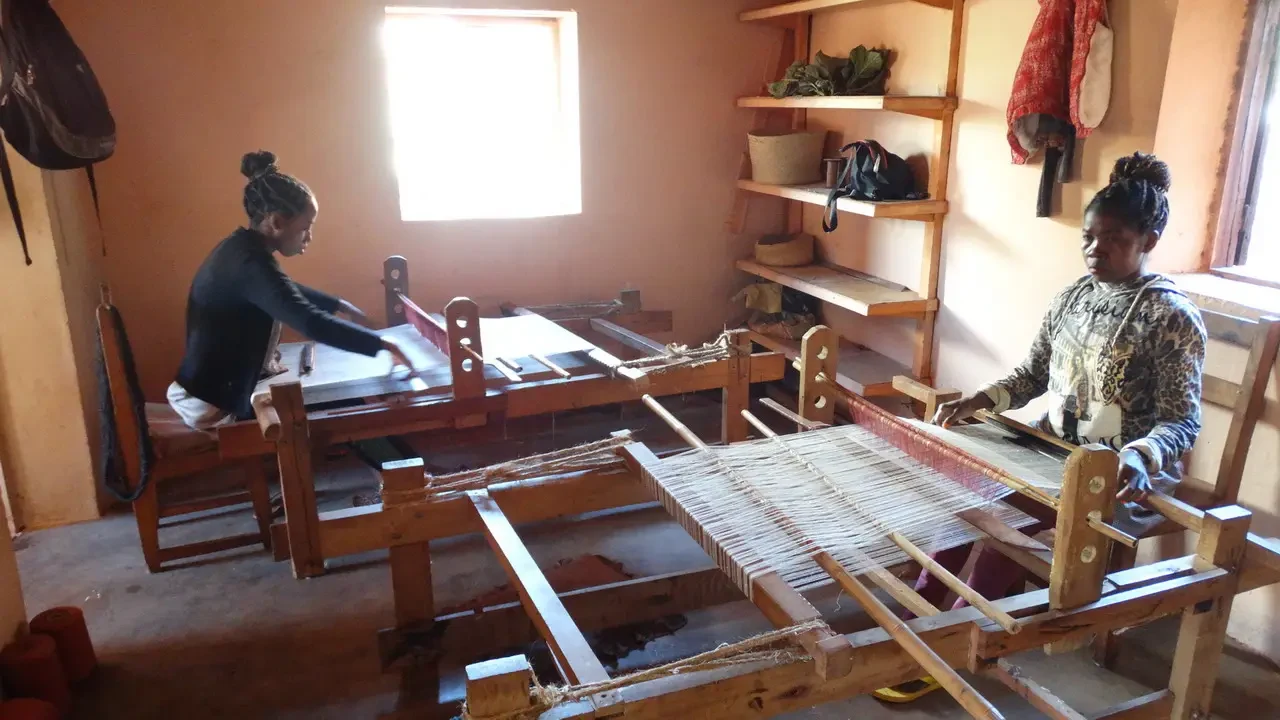 Due donne lavorano insieme su un telaio per la tessitura in un ambiente interno luminoso.