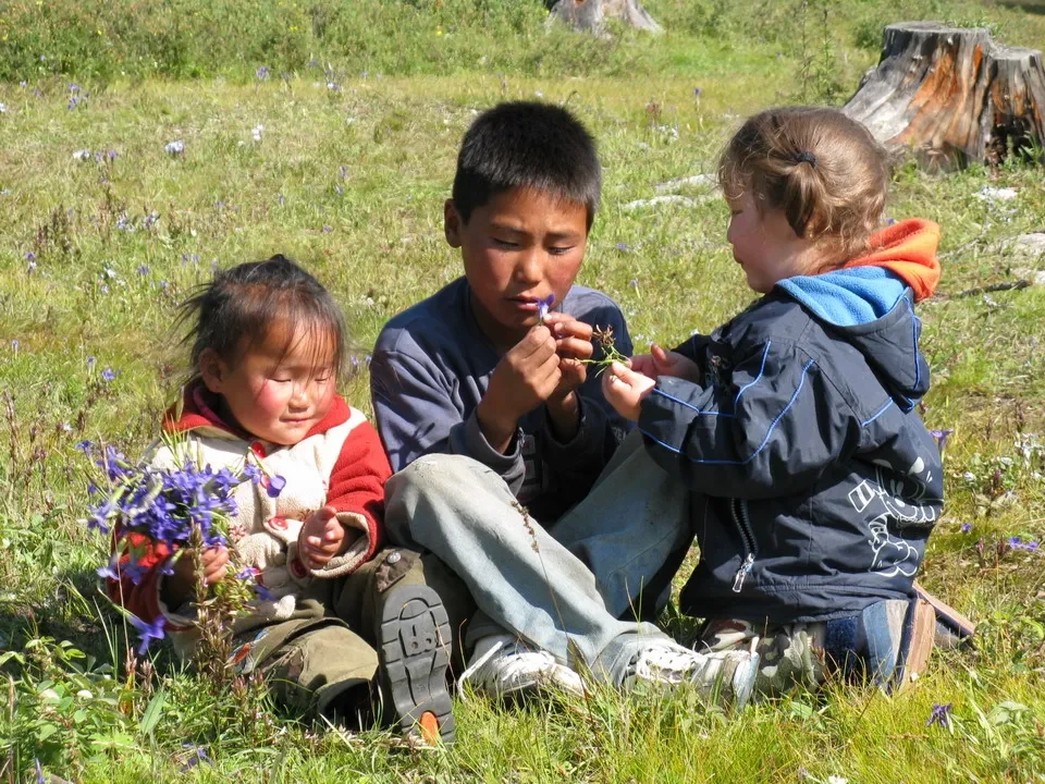 Tre bambini siedono a terra in un campo erboso con fiori.