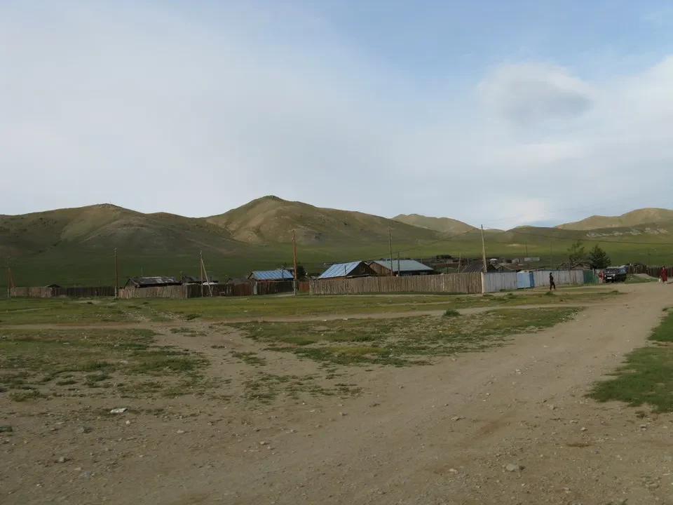 Paesaggio rurale della Mongolia con una stalla in legno e colline boscose sullo sfondo.