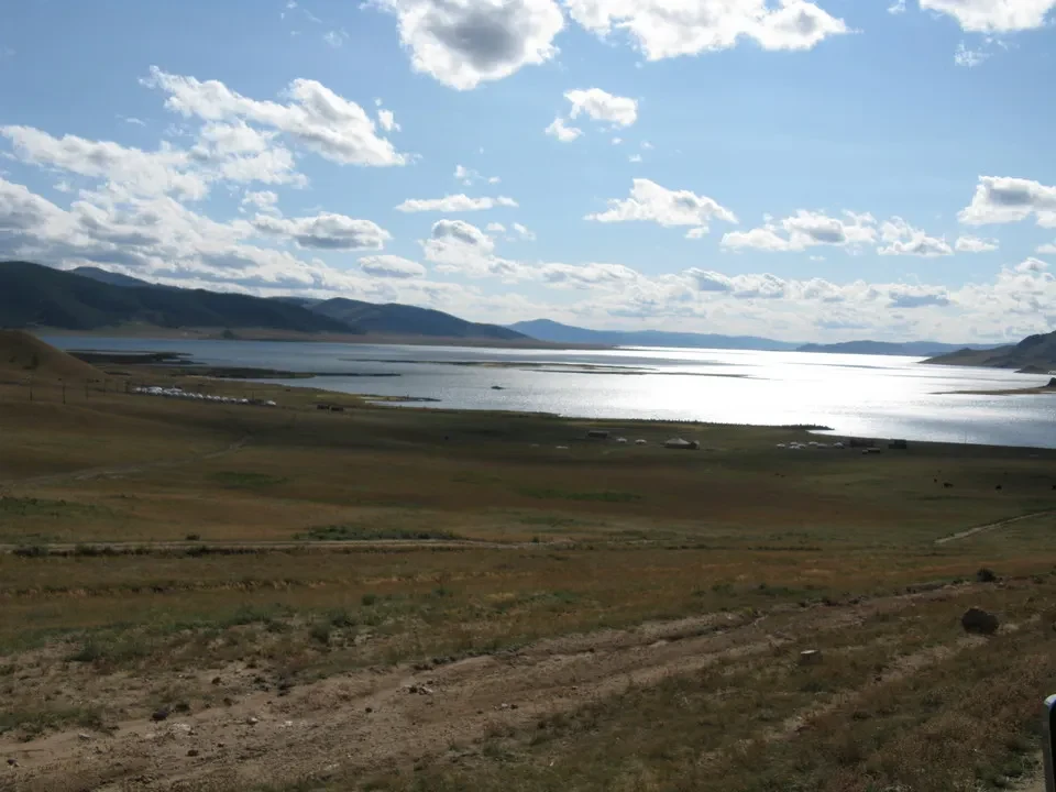 Un vasto paesaggio di praterie e un lago si estende sotto un cielo azzurro in Mongolia.