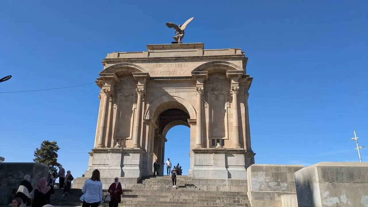 Arco trionfale monumentale in pietra con una statua in cima sotto un cielo azzurro.