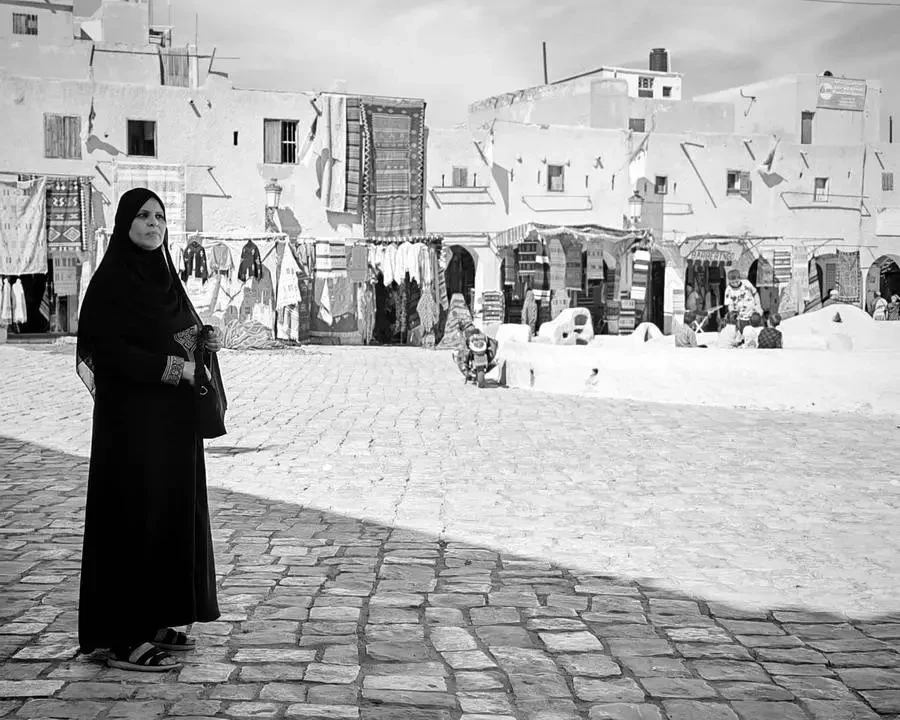 Una donna in abito scuro si trova davanti a edifici tradizionali in un contesto nordafricano.