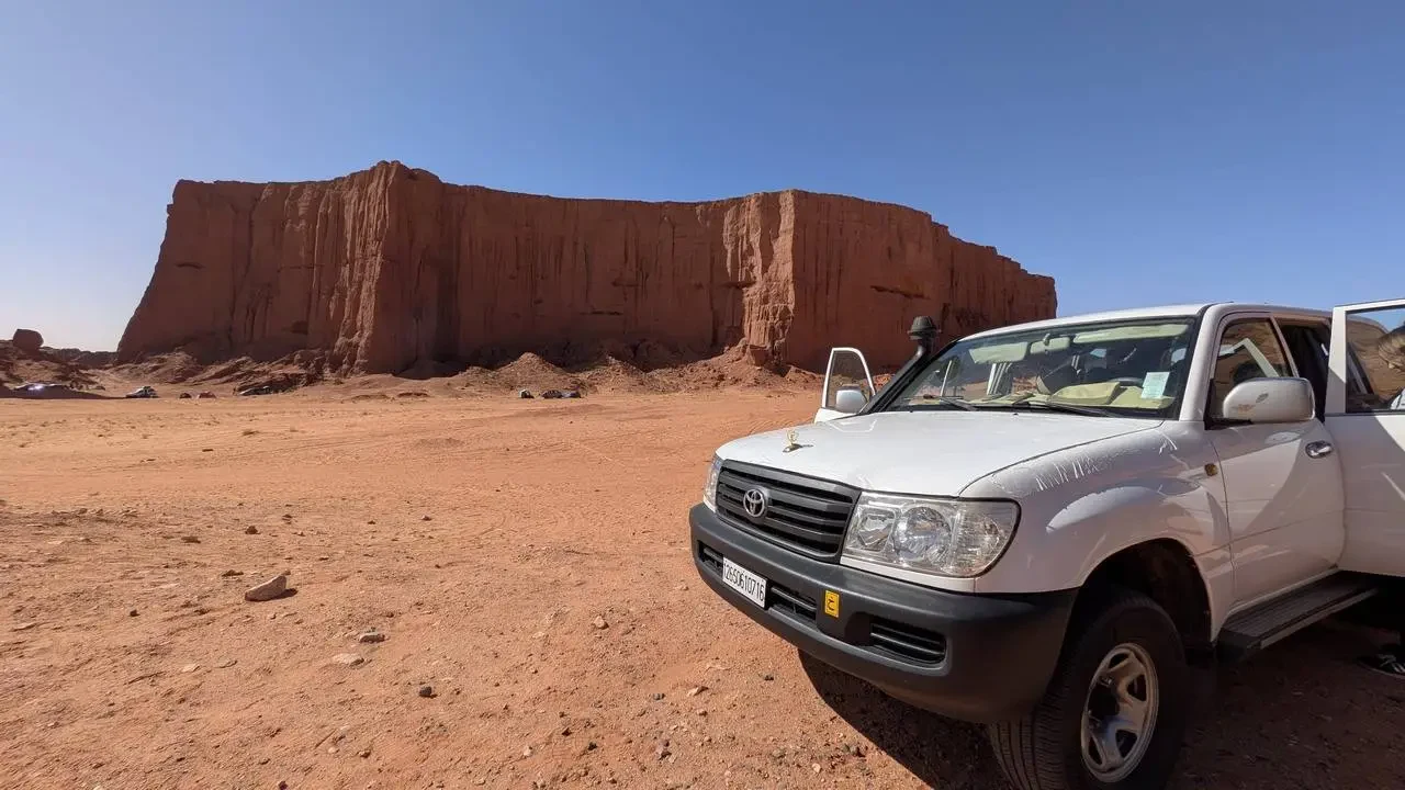 Un veicolo bianco è parcheggiato davanti a formazioni rocciose rosse nel deserto al tramonto.