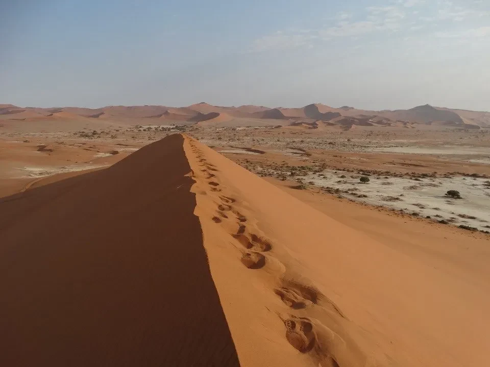 Una strada sterrata attraversa dune di sabbia arancione sotto un cielo chiaro.