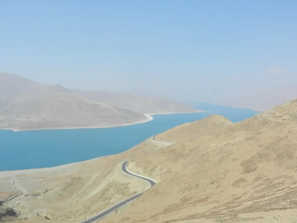 Paesaggio montano con un percorso sinuoso che costeggia un lago blu sotto il cielo aperto.