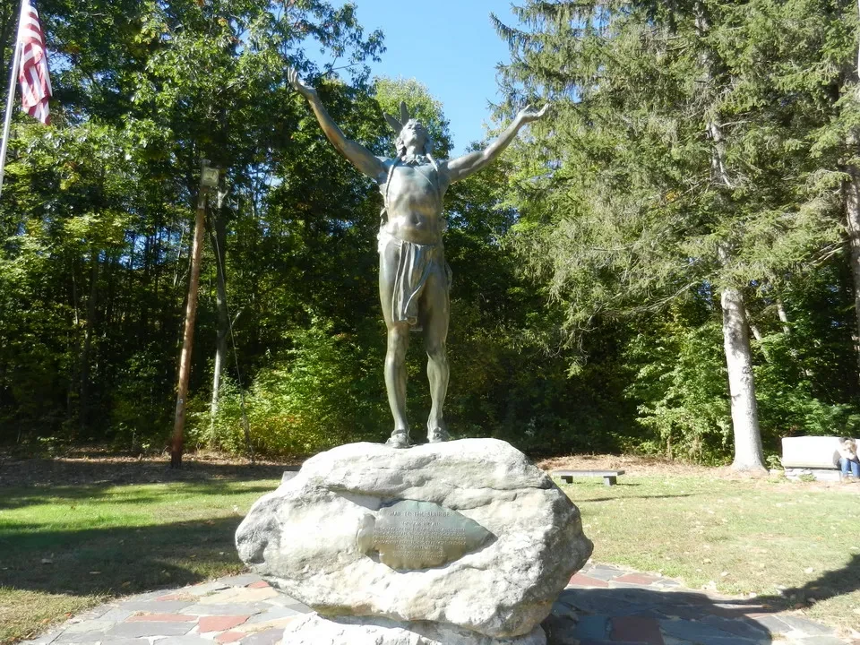 Statua di un uomo con le braccia aperte su una roccia in un ambiente boschivo.