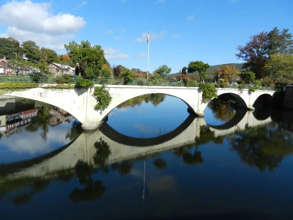 Un ponte ad arco in pietra attraversa l'acqua riflettendo gli alberi autunnali in un paesaggio del New England.