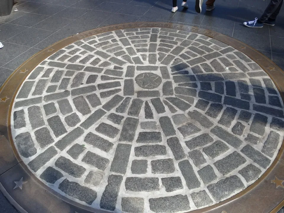Un motivo geometrico in pietra copre un'area pavimentata esterna dove le persone stanno camminando.