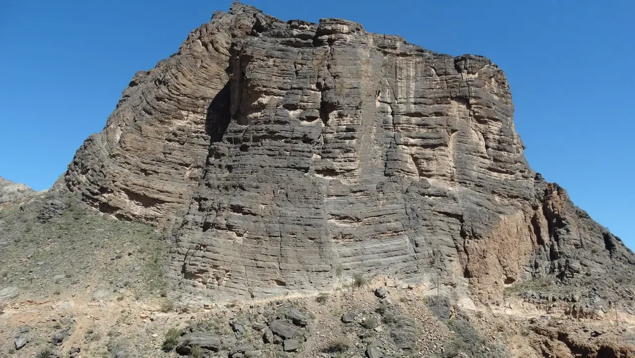 Una grande formazione rocciosa stratificata si erge dal terreno desertico arido sotto un cielo limpido.