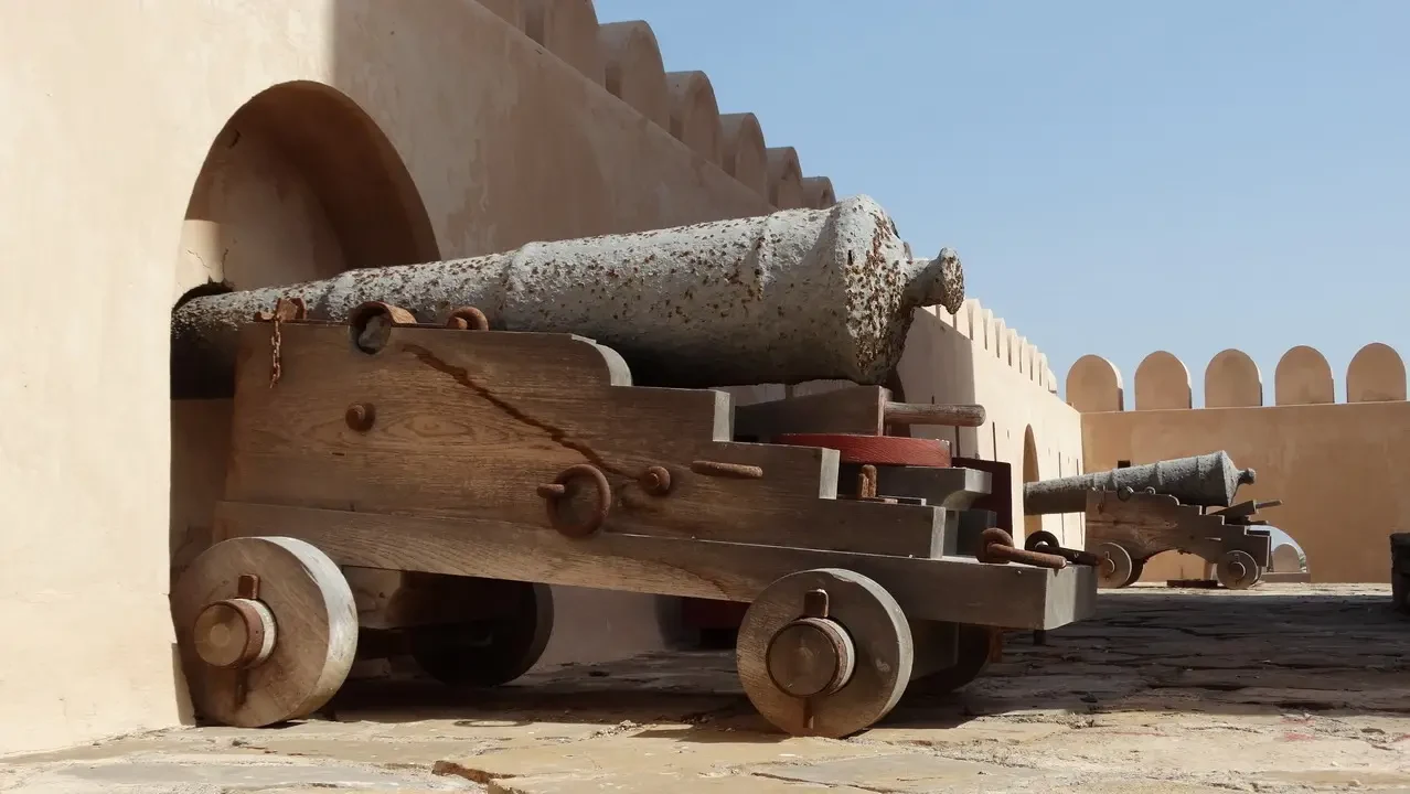 Un carro antico con un cannone è esposto lungo una fortificazione in pietra chiara.