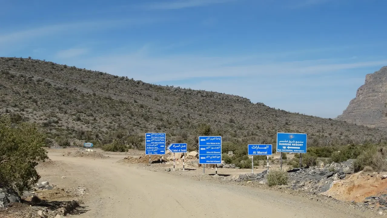 Una strada sterrata attraversa un paesaggio collinare arido con segnali stradali in Oman.