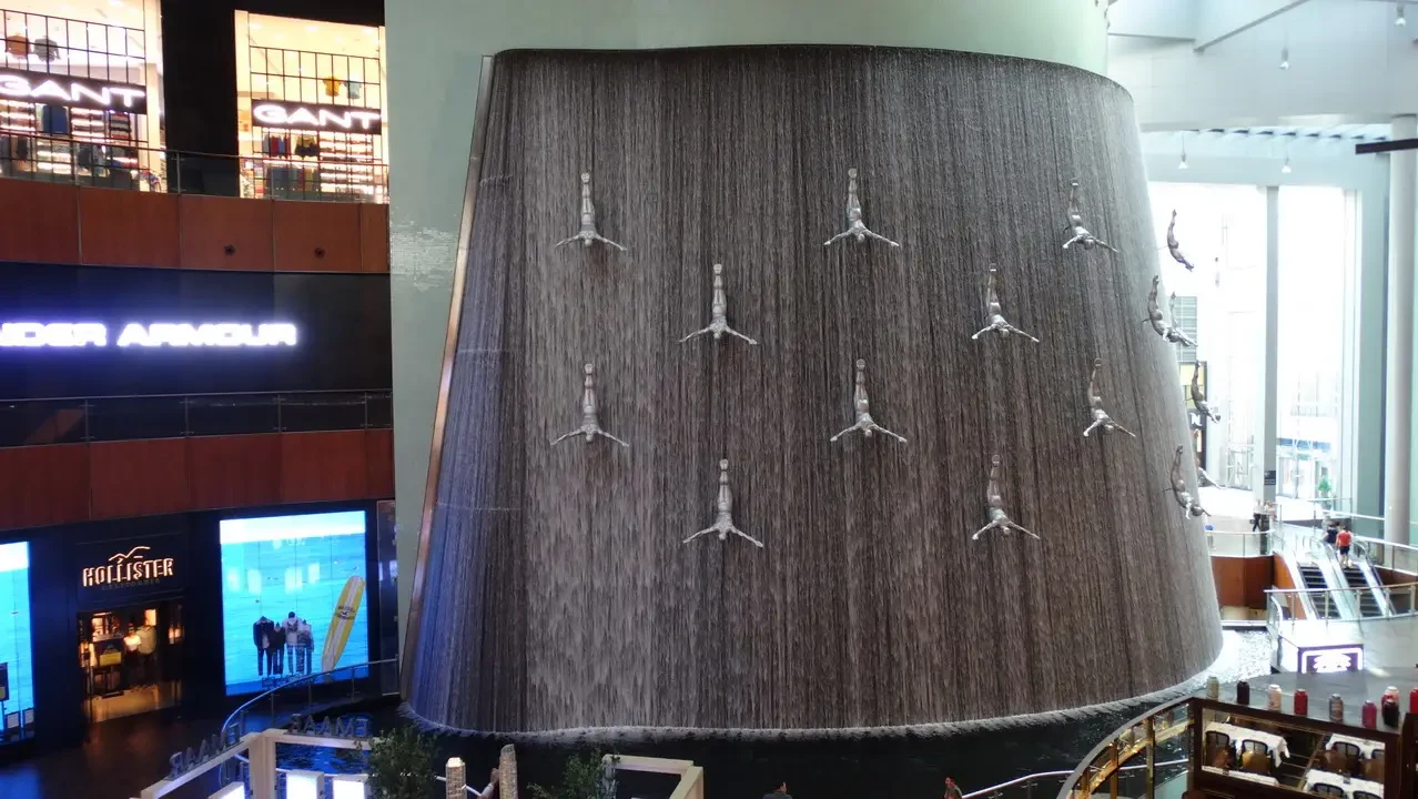 Una cascata d'acqua decorativa con forme a goccia cade su una grande parete verticale in un interno moderno.