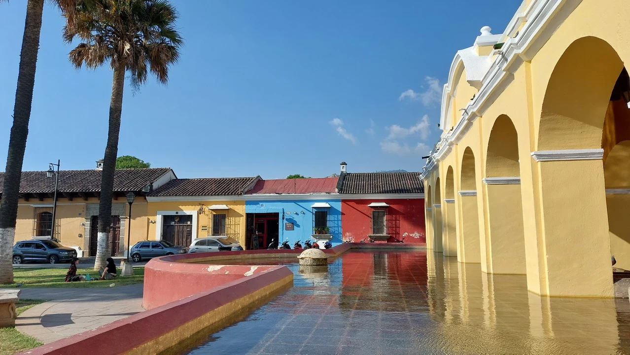 Una vista esterna di un edificio coloniale con archi gialli e una piazza bagnata in Antigua, Guatemala.