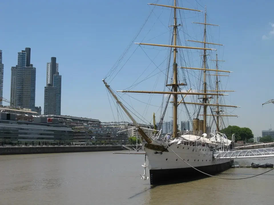 Una grande nave a vela ormeggiata in un fiume con edifici sullo sfondo.