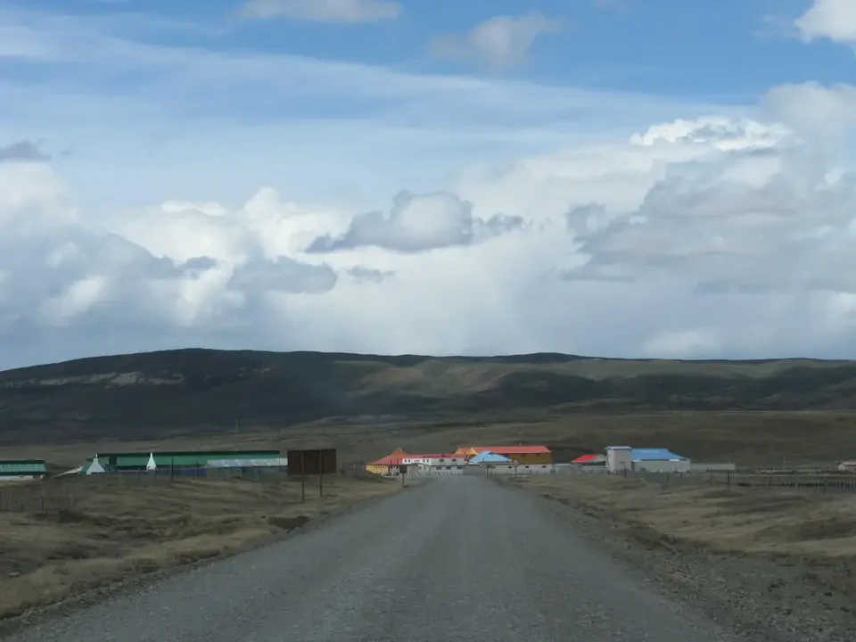 Una strada sterrata conduce attraverso un paesaggio aperto della Patagonia con edifici agricoli in lontananza.