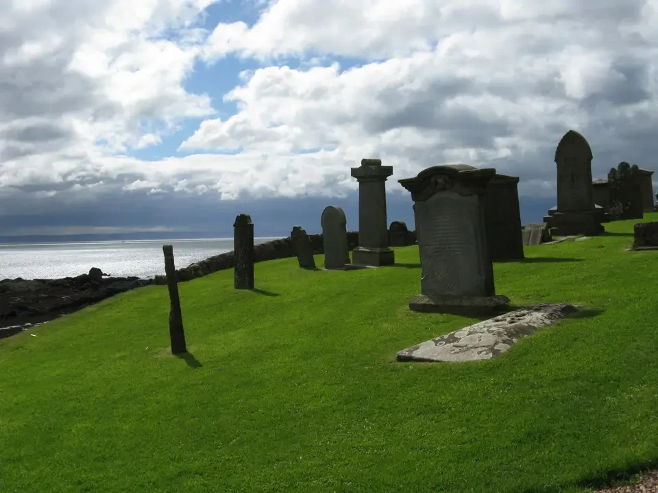 Cimitero con lapidi in primo piano su una collina erbosa vicino al mare.