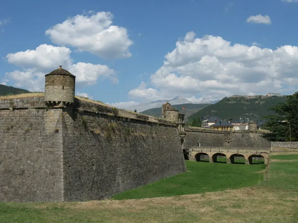 Mura di una fortezza in pietra con una torre visibile sotto un cielo nuvoloso.