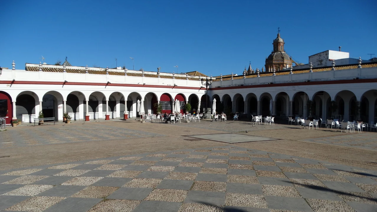 Una piazza pavimentata con portici architettonici e una chiesa in pietra sotto un cielo azzurro.