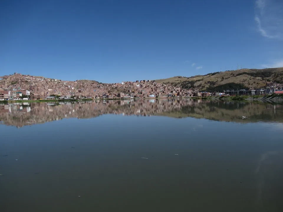 Riflesso delle colline e di un insediamento sull'acqua calma in un paesaggio andino.