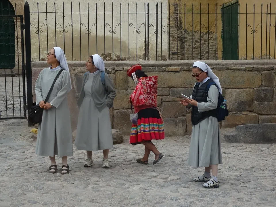 Quattro donne in abiti tradizionali camminano davanti a una recinzione in pietra.
