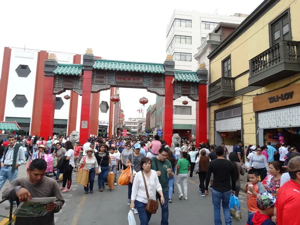 Una folla di persone attraversa un mercato o una strada con architetture tradizionali in Perù o Bolivia.