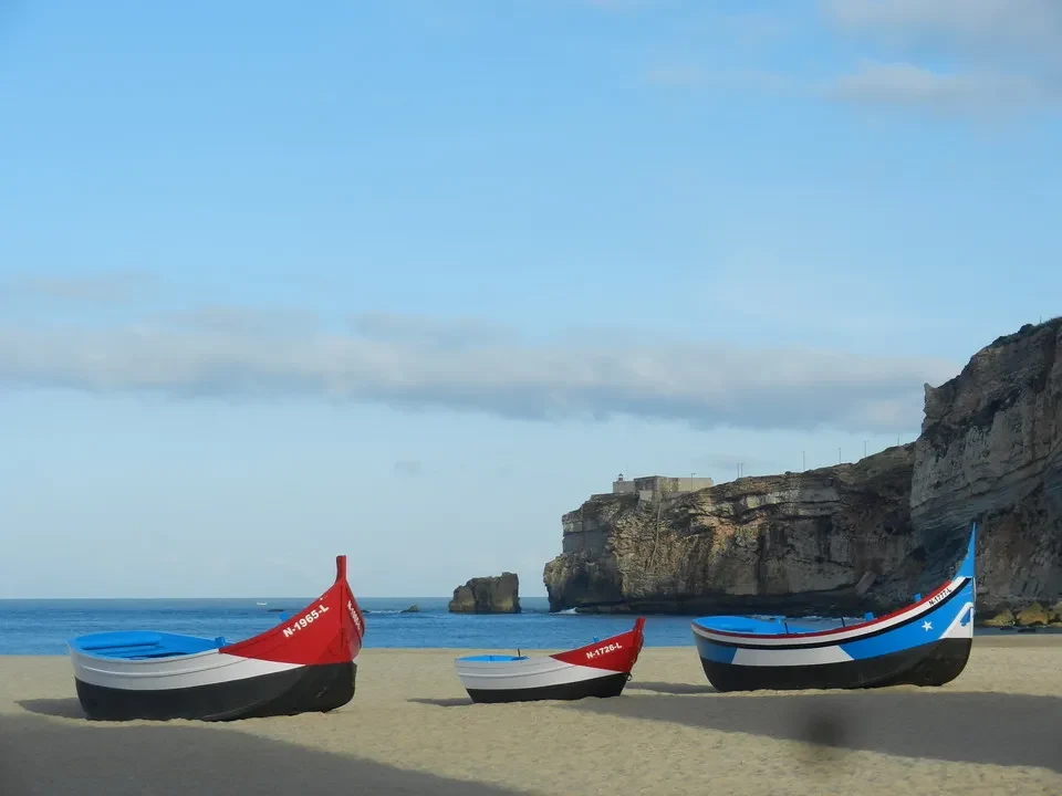 Barche colorate sono ormeggiate sulla spiaggia di Nazaré con scogliere rocciose sullo sfondo.