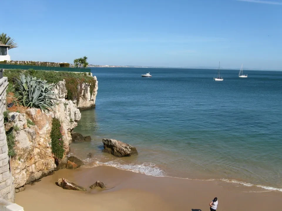 Spiaggia rocciosa e acque cristalline della costa portoghese con barche in lontananza.