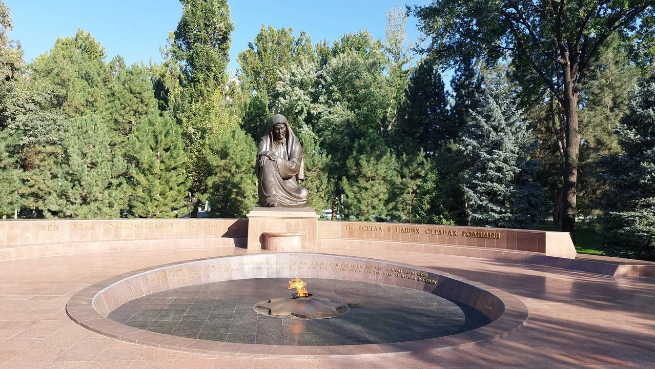 Una statua di bronzo in piedi è posizionata al centro di una piazza alberata con una fontana.